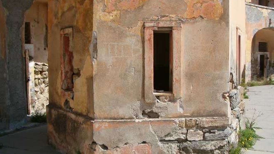 Facciata di un edificio antico e degradato, con intonaco scrostato e muratura in pietra a vista, e una finestra rettangolare senza vetro. L’edificio si affaccia su una strada lastricata, con archi e altri muri in rovina sullo sfondo.