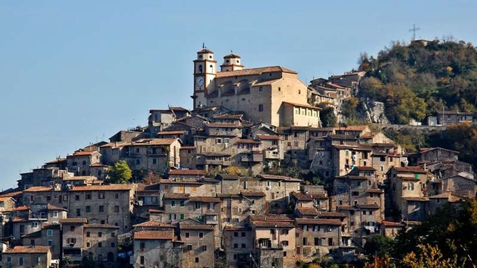 Borgo collinare con case in pietra addossate tra loro e tetti in coppi rossi, disposto a terrazze sul pendio. In cima si vede una grande chiesa con due campanili, con bosco e una croce su un rilievo sullo sfondo.