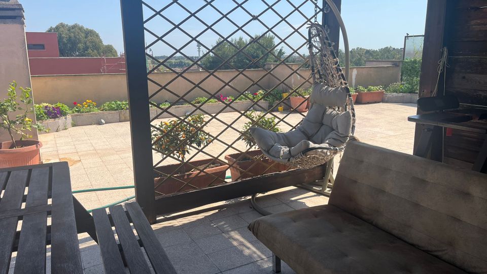 Terrazza sul tetto con pavimento piastrellato e area relax sotto una struttura in legno con grigliato, amaca a poltrona sospesa, divanetto e tavolino. Lungo il bordo ci sono fioriere e vasi con piante, con vista su edifici bassi e alberi sotto cielo sereno.