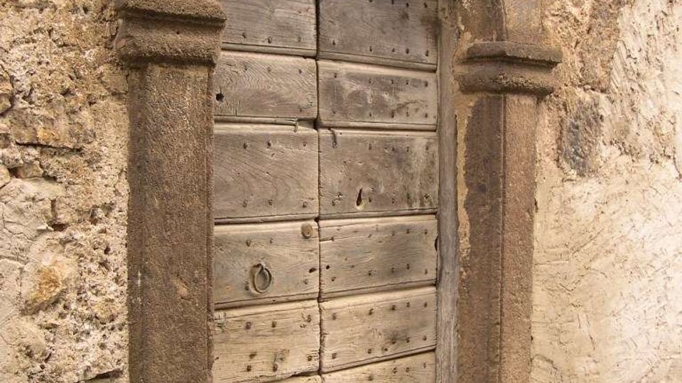 Porta antica in legno a due ante, con chiodature e battente in ferro, inserita in un arco in pietra scolpita su un muro di muratura grezza. Davanti alla soglia si vede un tratto di pavimentazione in ciottoli lungo una stretta strada.
