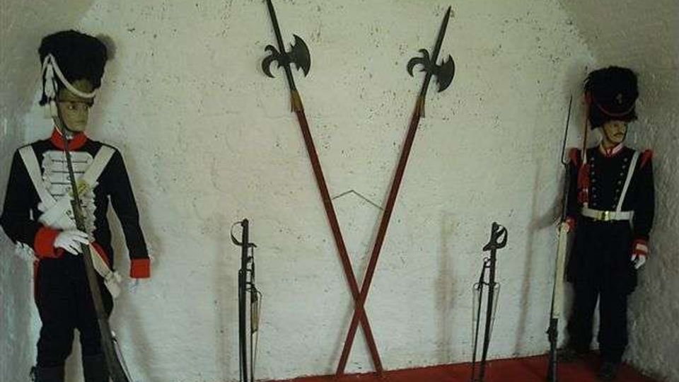 Interno di una piccola stanza con pareti bianche grezze e pavimento rosso, allestita come esposizione militare. Al centro sono appese due alabarde incrociate e ai lati sono disposti fucili e altre armi su supporti.