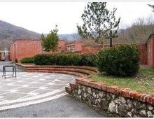 Cortile esterno pavimentato con disegni geometrici, delimitato da muretti in pietra e mattoni e aiuole con siepi e alcuni alberi. Sullo sfondo si vedono edifici bassi in mattoni rossi e colline boscose.