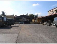 Ampio piazzale asfaltato di un’area industriale con capannoni e una tettoia metallica sullo sfondo, circondato da edifici bassi. Ai lati sono presenti diversi veicoli e attrezzature, con cielo sereno sopra.