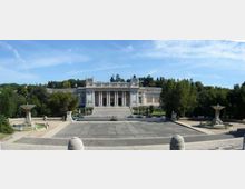 Vista panoramica di una grande piazza pavimentata con due fontane simmetriche ai lati, che conduce a un edificio monumentale in stile neoclassico con ampia scalinata e colonnato centrale. Sullo sfondo si vedono alberi e una collina verde sotto un cielo azzurro.