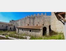 Cortile interno di una fortificazione in pietra con alte mura merlate e una piccola costruzione in pietra con tetto in coppi. Si vedono un arco d’ingresso, edifici annessi e aree erbose con muretti bassi sotto un cielo sereno.