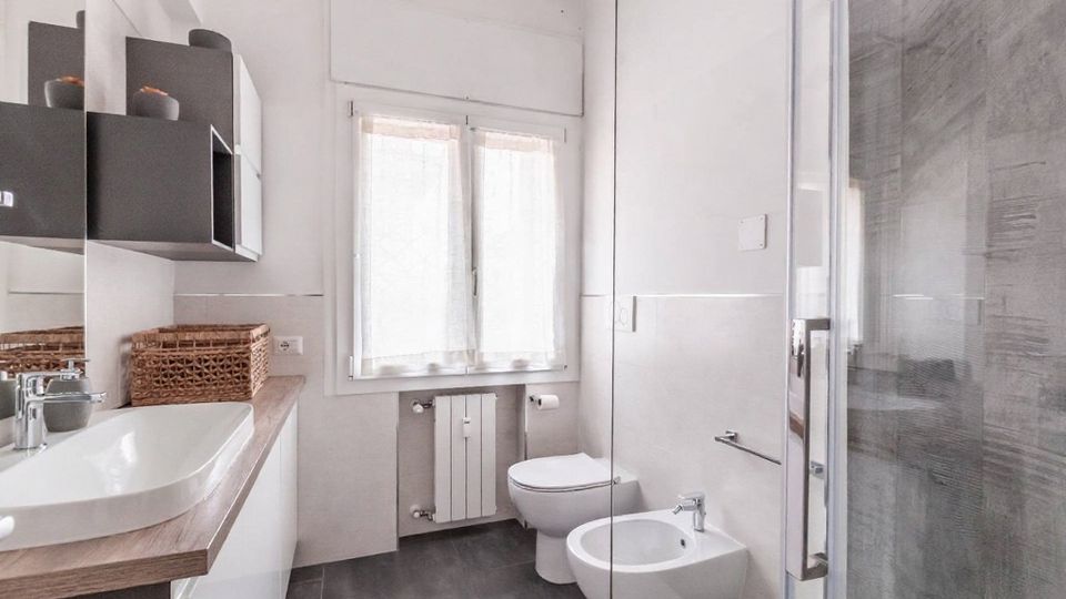 Bagno moderno e luminoso con piastrelle bianche e pavimento grigio scuro, mobile lavabo con piano in legno e pensili scuri. Sul fondo c’è una finestra con tenda e radiatore, mentre a destra si vedono WC, bidet e una doccia con box in vetro.