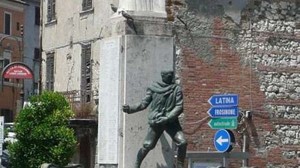 Monumento in piazza con basamento in pietra e colonna alta sormontata da una statua di angelo; alla base è presente una statua in bronzo e due lapidi commemorative con iscrizioni. Sullo sfondo si vedono edifici storici con intonaco consumato e cartelli stradali su un incrocio urbano.