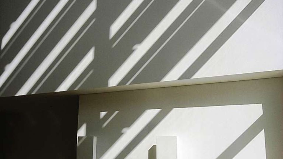 Atrio interno moderno con pareti bianche e pavimento in grandi lastre, attraversato da lunghe ombre diagonali create da una vetrata o lucernario. Sul lato sinistro si intravedono sedute e un’area più in ombra.
