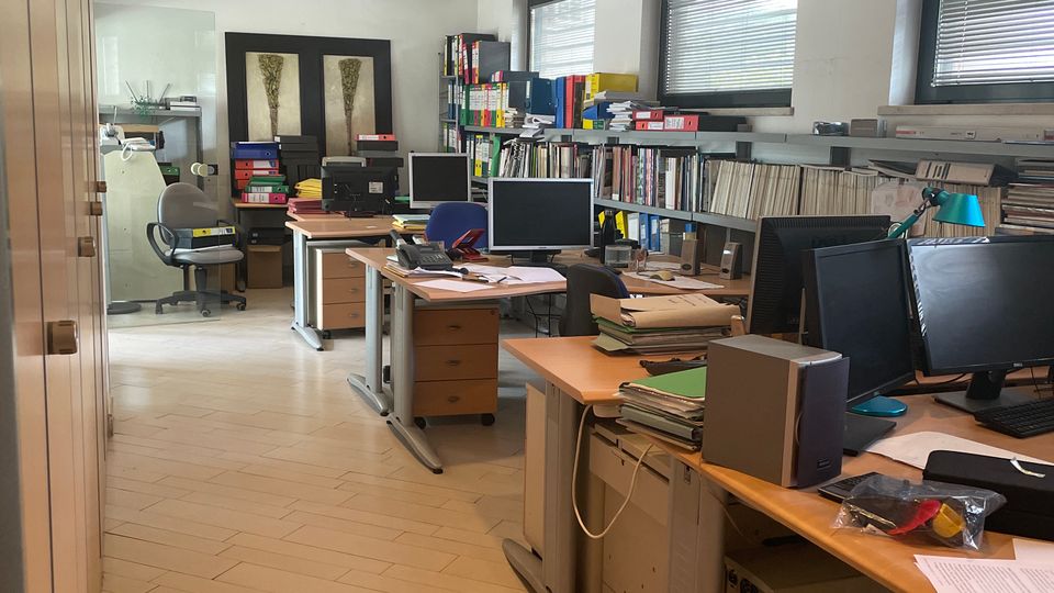 Interno di un ufficio condiviso con più scrivanie in fila, computer e pile di documenti, affiancato da scaffali pieni di raccoglitori e libri. Pareti bianche con finestre dotate di veneziane, pavimento in piastrelle chiare e illuminazione a faretti incassati nel soffitto.