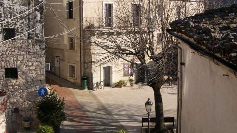 Piccola piazza di un borgo con edifici in pietra e intonaco dai tetti in coppi, affacciati su una strada lastricata. Al centro si vedono un albero spoglio, panchine, un lampione e alcune fioriere, con colline boscose sullo sfondo.