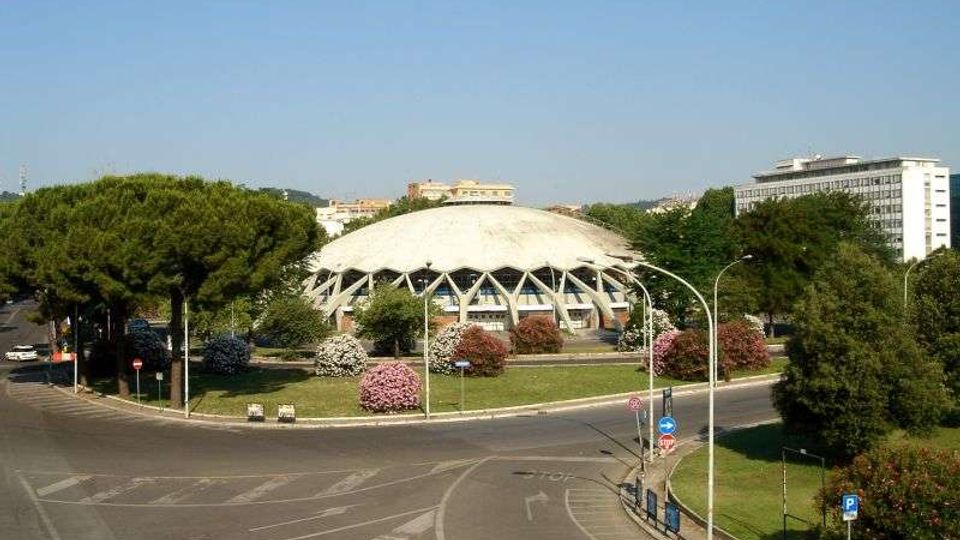 Edificio moderno a pianta circolare con grande cupola bianca e struttura esterna a travi, situato al centro di un’area verde con alberi e aiuole fiorite. In primo piano si vede una rotatoria stradale, con palazzi e vegetazione sullo sfondo.
