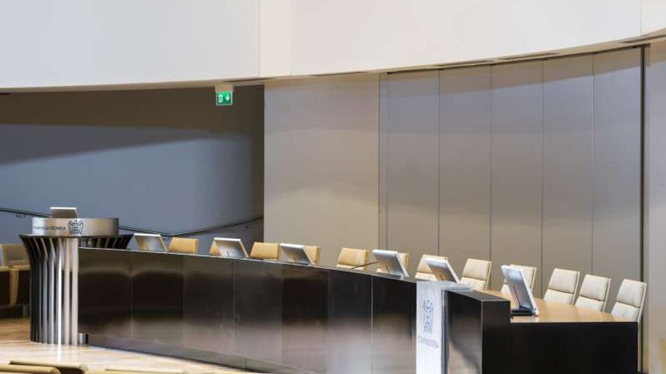 Sala conferenze moderna con un lungo banco curvo in metallo e legno, dotato di microfoni e schermi, e file di sedute in platea in primo piano. Sullo sfondo si vedono pareti lisce e pannelli chiari con un’uscita di emergenza segnalata.