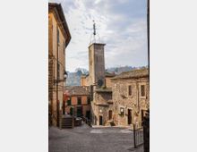 Strada in discesa in un borgo storico con case in pietra e intonaco, tetti in coppi e lampioni in ferro battuto. Al centro si vede una torre quadrata con struttura metallica in sommità, sullo sfondo colline e cielo nuvoloso.