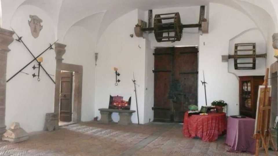 Interno di una sala storica con soffitto a volte bianche e pavimento in mattoni a spina di pesce, con grande portone in legno sul fondo. Alle pareti sono esposte armi e attrezzi antichi, con una piccola panca in pietra e alcuni tavoli coperti da teli.