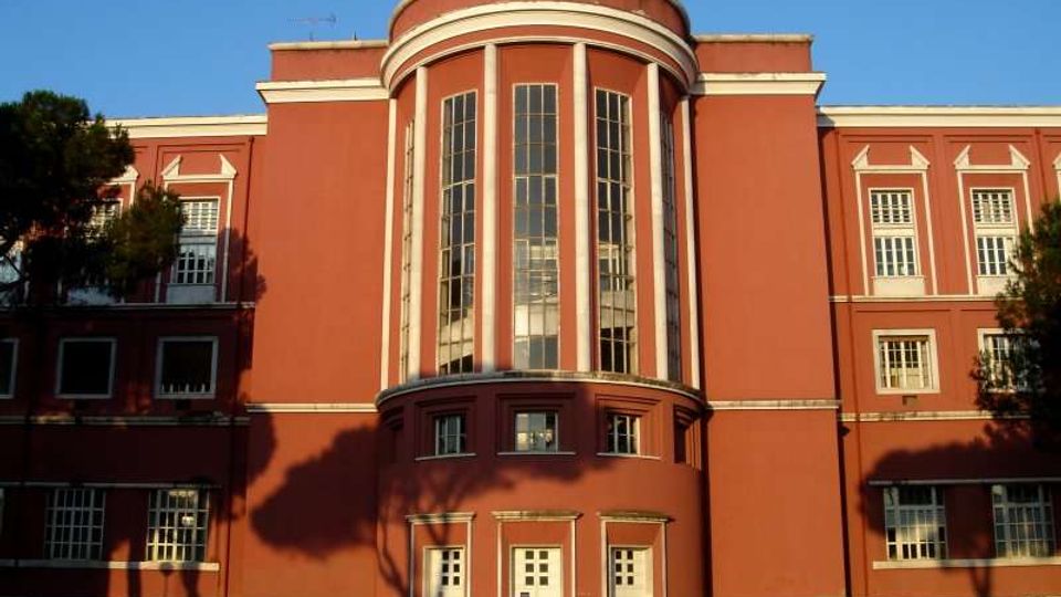 Facciata di un grande edificio in stile razionalista, intonacato in rosso con cornici bianche e un volume centrale semicircolare con alte finestre verticali. Ai lati si vedono file di finestre rettangolari e un’area esterna pavimentata davanti all’ingresso.