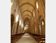 Interno di una chiesa in pietra con lunga navata centrale, arcate su colonne e soffitto a volte ogivali. File di panche in legno conducono verso l’abside, illuminata da una finestra a rosone e da strette finestre laterali.