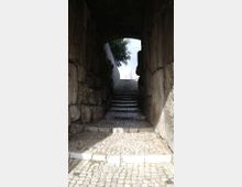 Passaggio in pietra con volta ad arco e pavimentazione in ciottoli, che conduce a una scalinata ripida. L’uscita in alto è illuminata e incornicia cielo e vegetazione sullo sfondo.