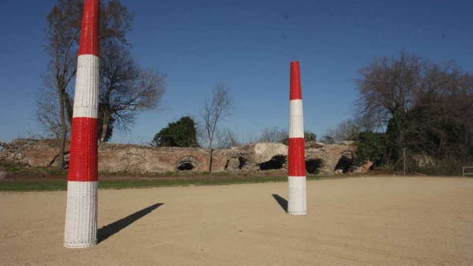 Campo aperto con terreno sabbioso e due alti pali cilindrici a bande rosse e bianche. Sullo sfondo si vede un muro in pietra con archi e alberi spogli sotto un cielo sereno.
