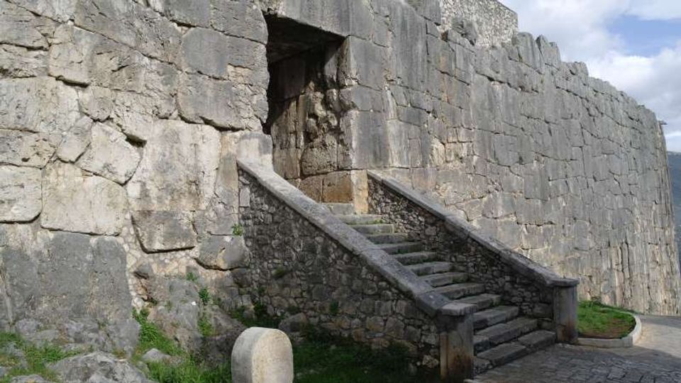 Muro di fortificazione in grandi blocchi di pietra con un’apertura in alto raggiungibile tramite una scalinata in pietra con muretti laterali. In primo piano si vede un tratto di pavimentazione e una piccola area erbosa ai piedi della muratura.