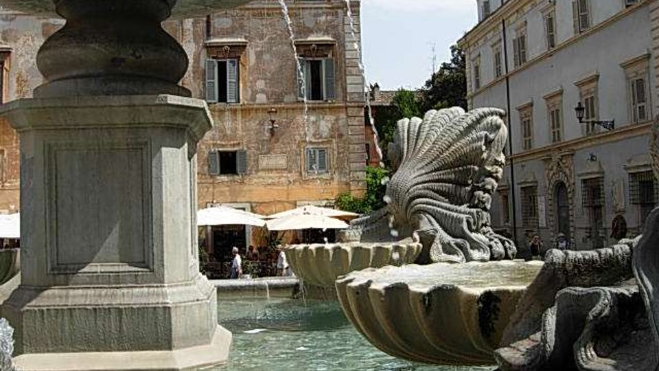 Fontana monumentale in una piazza urbana, con grande vasca d’acqua e sculture in pietra a forma di conchiglia e creature marine. Sullo sfondo si vedono palazzi storici con facciate intonacate e finestre, oltre a ombrelloni di un’area ristoro.