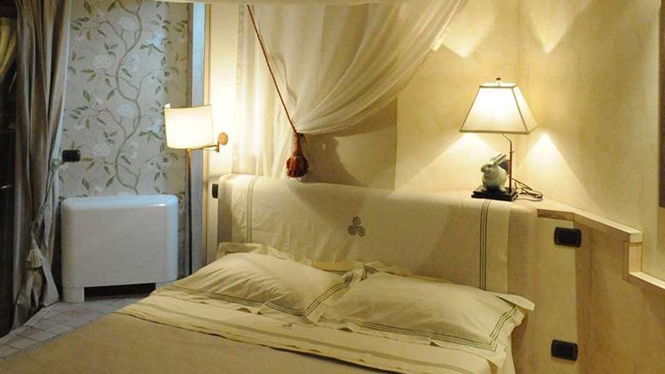 Camera da letto in stile rustico con letto matrimoniale, biancheria chiara e baldacchino in tessuto appeso al soffitto. Ai lati ci sono lampade da comodino su una testiera in muratura, con tende e parete decorata sullo sfondo.