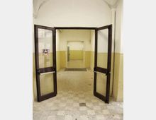 Ingresso interno con doppia porta a battente in legno scuro e vetro, aperta su un corridoio con pareti bianche e fascia inferiore giallo pallido. Pavimento in piastrelle grigie a scacchiera e soffitto con volte ad arco.