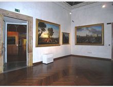 Sala di museo con pavimento in parquet scuro e pareti bianche, con grandi dipinti incorniciati appesi alle pareti e illuminazione a soffitto. A sinistra si vede un’apertura con cornice in pietra che conduce a un’altra galleria.