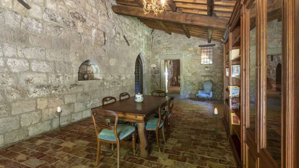Sala interna in stile rustico con pareti in pietra a vista, soffitto con travi in legno e pavimento in mattoni. Al centro c’è un tavolo da pranzo con sedie, mentre lungo un lato si vede una grande libreria in legno e sul fondo archi e una finestra che illuminano il corridoio.