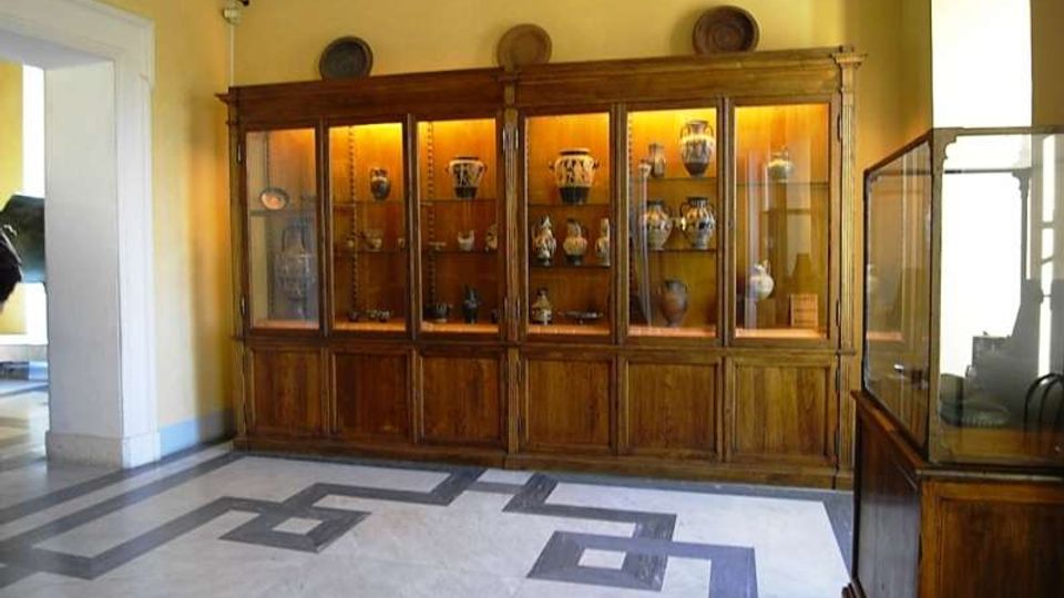 Interno di sala museale con pareti gialle e pavimento in marmo a motivi geometrici, dominata da una grande vetrina in legno illuminata che espone vasi e ceramiche. A destra è visibile un’altra teca in vetro, mentre a sinistra si apre un passaggio verso un ambiente adiacente.