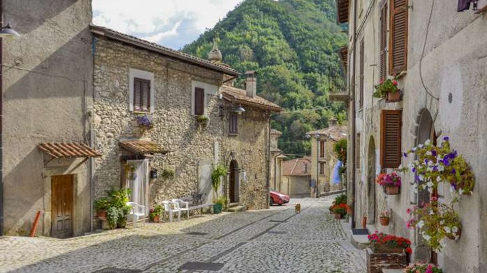 Strada acciottolata in un borgo collinare, fiancheggiata da case in pietra e intonaco con persiane in legno, archi e vasi di fiori sui davanzali. Sullo sfondo si vedono un campanile e una collina boscosa che domina l’abitato.