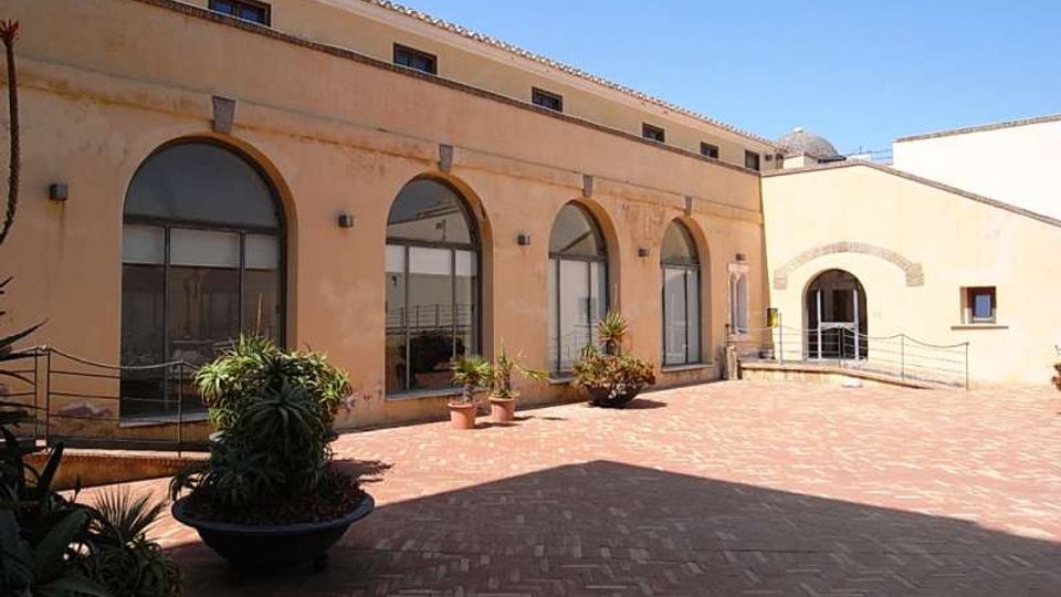 Cortile lastricato in mattoni con vasi di piante, affiancato da un edificio intonacato color ocra con grandi finestre ad arco. Sullo sfondo si vede un ingresso ad arco in un’ala adiacente e un cielo sereno sopra i tetti in coppi.