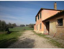 Casa di campagna a due piani intonacata color rosa con persiane verdi e tetto in coppi, affiancata da un annesso più basso con finestra. Davanti si estende un vialetto di ghiaia che conduce a campi verdi con alberi sparsi sotto un cielo sereno.