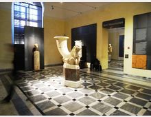Sala museale con pavimento a scacchi in marmo e pareti gialle, con grandi pannelli espositivi neri e una finestra ad arco alta. Al centro è esposto un grande reperto scultoreo in pietra su piedistallo, con altre opere e testi informativi lungo le pareti e un passaggio verso un’altra sala.