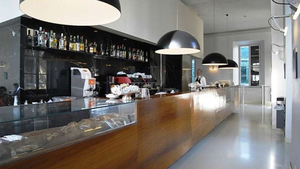 Interno di un bar moderno con lungo bancone in legno e vetrina, parete nera con scaffali di bottiglie e grandi lampade a sospensione a cupola. Pavimento chiaro lucido, soffitto alto e grandi finestre sul fondo che illuminano l’ambiente.