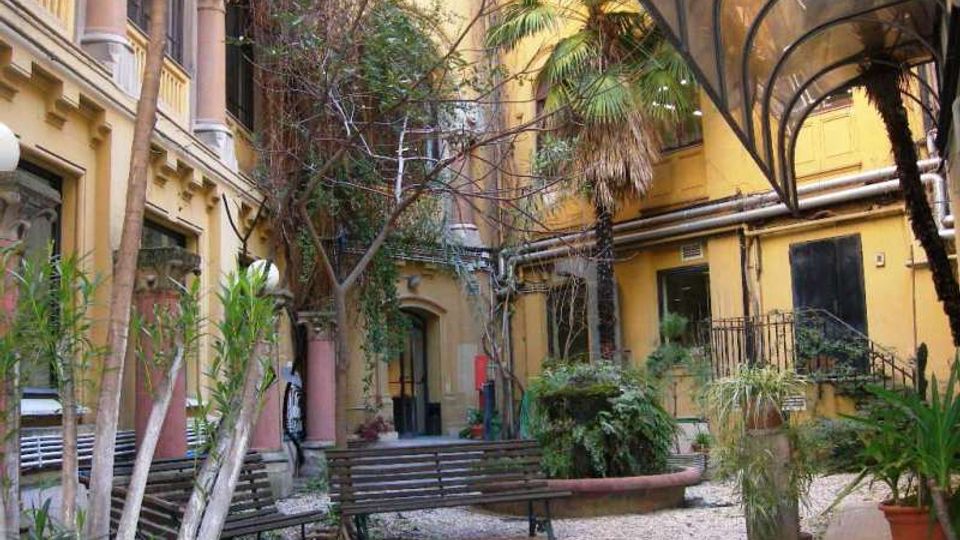 Cortile interno di un edificio con facciate gialle e balconi, pavimentato in ghiaia e arredato con panchine. Sono presenti molte piante e alberi in vaso e un’ampia tettoia trasparente ad arco lungo il lato destro.