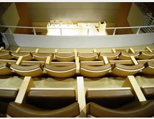 Interno di un’aula magna o auditorium con file di sedute imbottite color beige disposte in gradoni e rivolte verso un palco. In fondo si vede un lungo tavolo con sedie e un leggio, separati da una balaustra chiara.