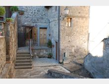 Cortile in un borgo con case in pietra grezza, piccola scalinata e portoncino con balconcino in ferro. Sul muro sono visibili una lanterna, finestre con persiane e alcuni vasi di fiori.