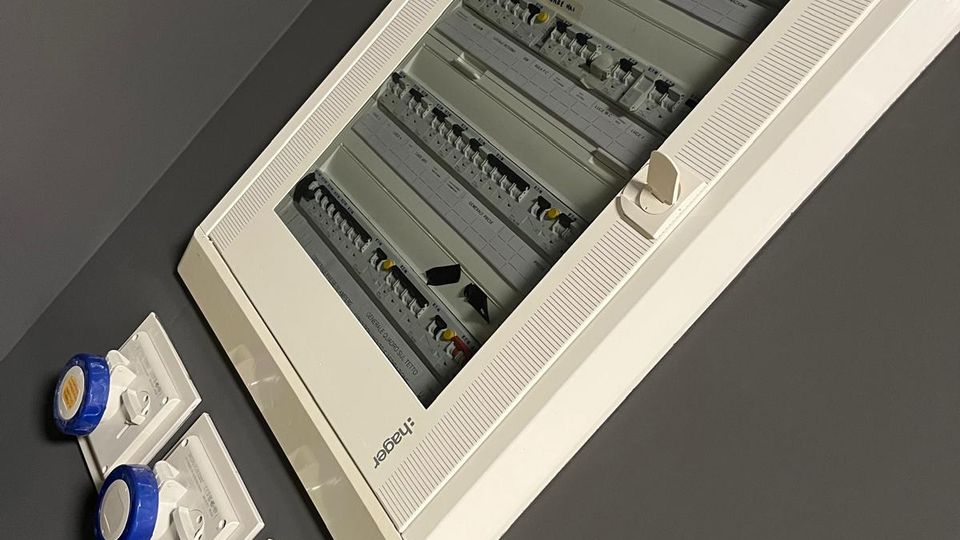 Quadro elettrico da parete con sportello trasparente, contenente interruttori e moduli su guide DIN, montato su una parete grigia. Accanto sono visibili tre prese industriali blu su placche bianche.