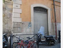 Facciata di un edificio urbano con intonaco ocra e basamento in pietra, caratterizzata da una grande porta ad arco con cornice chiara e grata decorativa. Davanti, su strada in sampietrini, sono parcheggiati scooter e biciclette vicino a una ringhiera metallica e a paletti.
