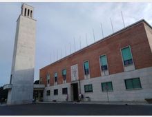 Edificio civico a due piani con facciata in mattoni rossi e basamento in pietra chiara, con ingresso centrale e finestre rettangolari. A sinistra si innalza una torre campanaria moderna in cemento chiaro, in una piazza aperta sotto un cielo nuvoloso.