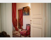 Angolo interno in stile classico con poltrona in legno imbottita a righe rosse, specchio alto con tende a righe rosse e tappeto. Sulla parete bianca sono visibili un quadro e una lampada a parete, con una porta bianca in primo piano.