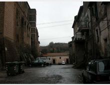 Piazzale lastricato in pietra tra edifici in muratura antica, con facciate scure, finestre e balconi in ferro. Sullo sfondo si vede un edificio basso con tetto in tegole e una collina alberata, con auto parcheggiate e cassonetti lungo i lati.