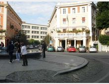 Piazza urbana con una fontana circolare in primo piano e una strada curva lastricata che la costeggia. Sullo sfondo si vedono palazzi a più piani con facciate chiare e balconi, auto parcheggiate lungo il bordo e alberi sul lato destro.