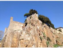 Rovine di una fortificazione in pietra con merli, arroccata sulla sommità di una rupe rocciosa. Sulla cima e lungo il crinale si vedono alberi e vegetazione, con cielo sereno sullo sfondo.