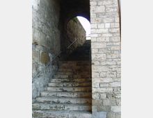 Scalinata in pietra consumata che sale all’interno di un passaggio ad arco, tra alti muri di muratura grezza. Corrimani metallici corrono lungo i lati e in cima si intravede luce naturale dall’apertura superiore.