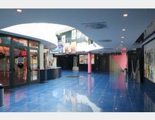 Atrio interno di un cinema o complesso commerciale con pavimento lucido in piastrelle blu, soffitto bianco curvo con faretti e lucernari. Ai lati si vedono ingressi vetrati, colonne rosa, biglietteria e pareti con poster e murales.