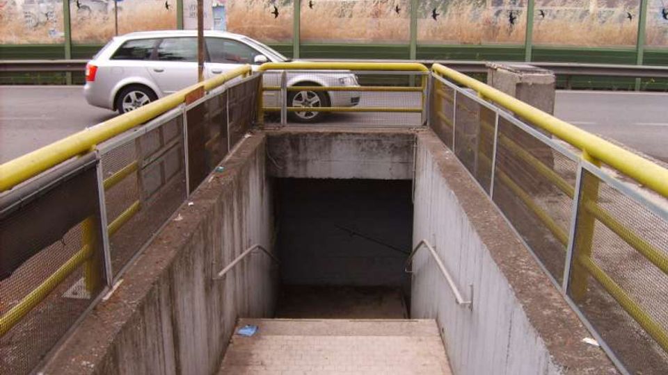 Ingresso a un sottopassaggio pedonale con scala in cemento che scende sotto la strada, affiancata da corrimani e parapetti con ringhiere gialle. Sullo sfondo si vede la carreggiata con un’auto e una barriera fonoassorbente verde lungo la strada.