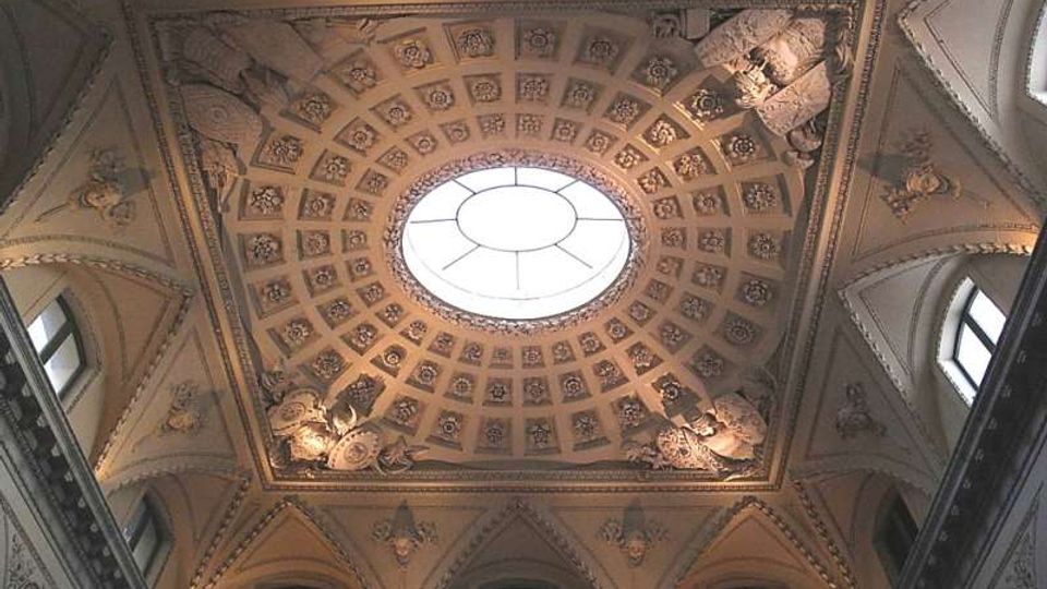 Soffitto interno di un edificio storico con grande lucernario ovale centrale, circondato da cassettoni decorati e rilievi scultorei agli angoli. Lungo il perimetro si vedono archi a sesto acuto e finestre superiori incorniciate da stucchi ornamentali.