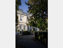 Facciata di una villa storica color crema con finestre incorniciate e balconata in sommità, parzialmente coperta da grandi alberi. In primo piano un vialetto nel giardino con siepi, vasi e una piccola statua vicino all’ingresso con portico.