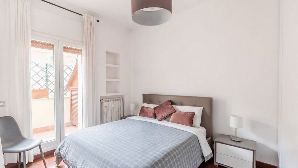 Camera da letto luminosa con letto matrimoniale, comodini con lampade e pavimento in legno; grandi porte finestre con tende bianche si aprono su un piccolo balcone. Pareti bianche con nicchie a muro, radiatore e lampadario a soffitto.